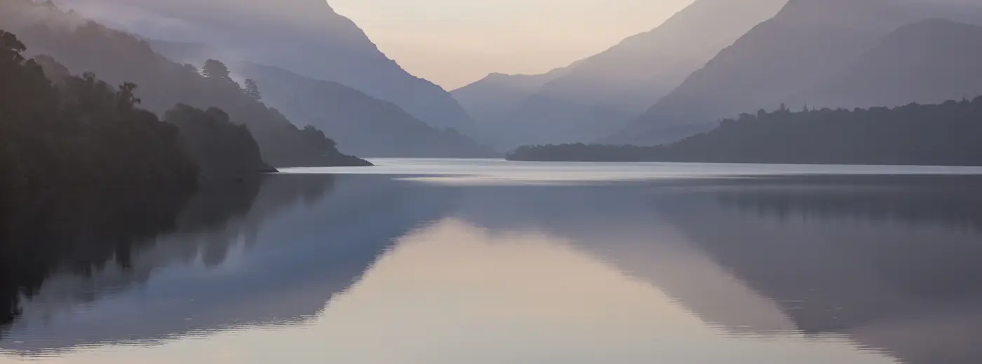 A still lake in a Welsh valley at first light, low mist lying on the water and the hills fading into one another behind it.